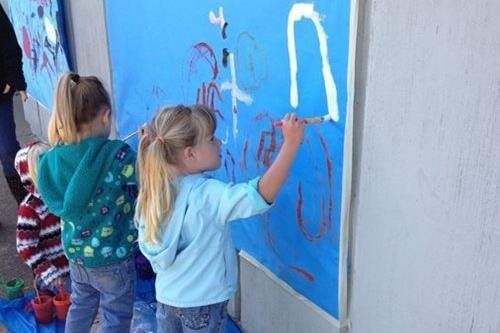 Two children paint colorful shapes on a blue wall outdoors