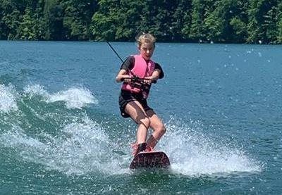 Person wakesurfs on a lake, wearing a pink life jacket with a wooded shoreline in the background