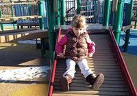 Child sitting on a playground slide, wearing a pink jacket and white pants.