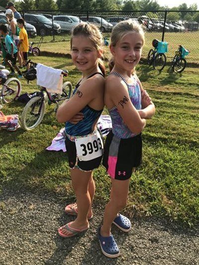 Two children standing back to back at a bike race, smiling with numbered bibs on and bicycles behind them.