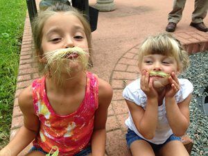 Two children sitting on steps, making funny faces with grass under their noses and mouths.