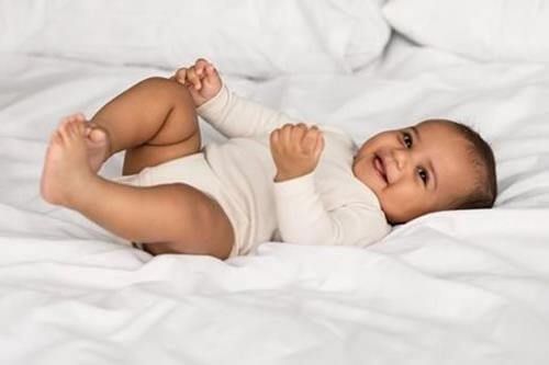 Smiling baby in a white outfit lying on a white blanket, kicking legs and holding hands up