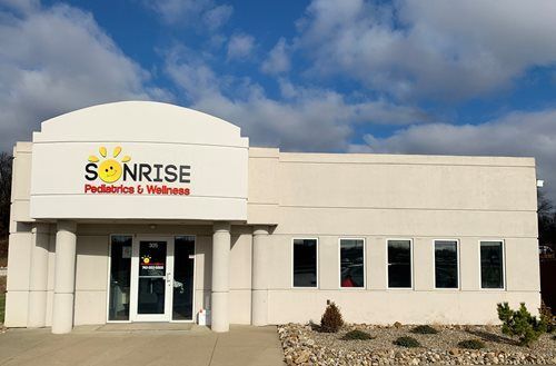 Modern building with a cross-shaped design, glass windows, and a sign. Palm trees and parked cars are in front.