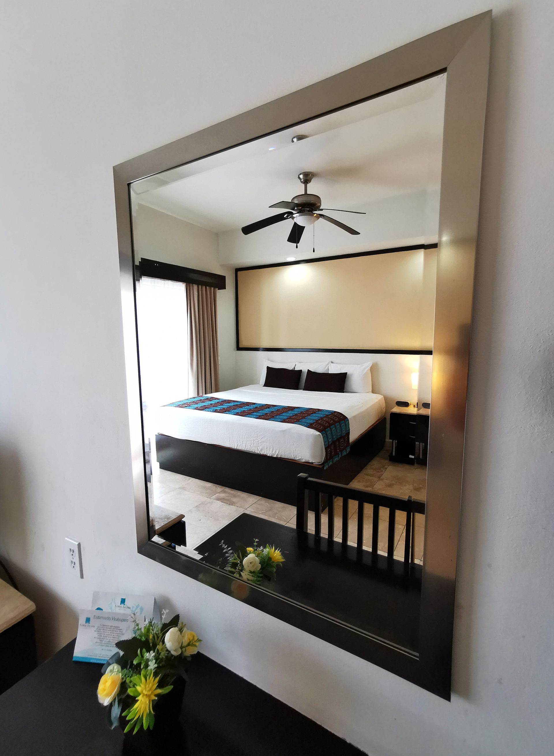 A bedroom with a king size bed and a ceiling fan is reflected in a mirror.