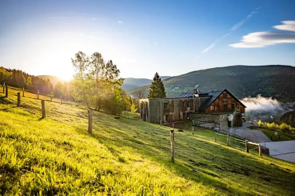 Pendio verdeggiante con un caratteristico hotel, un albero e una recinzione, sullo sfondo un sole splendente e delle montagne