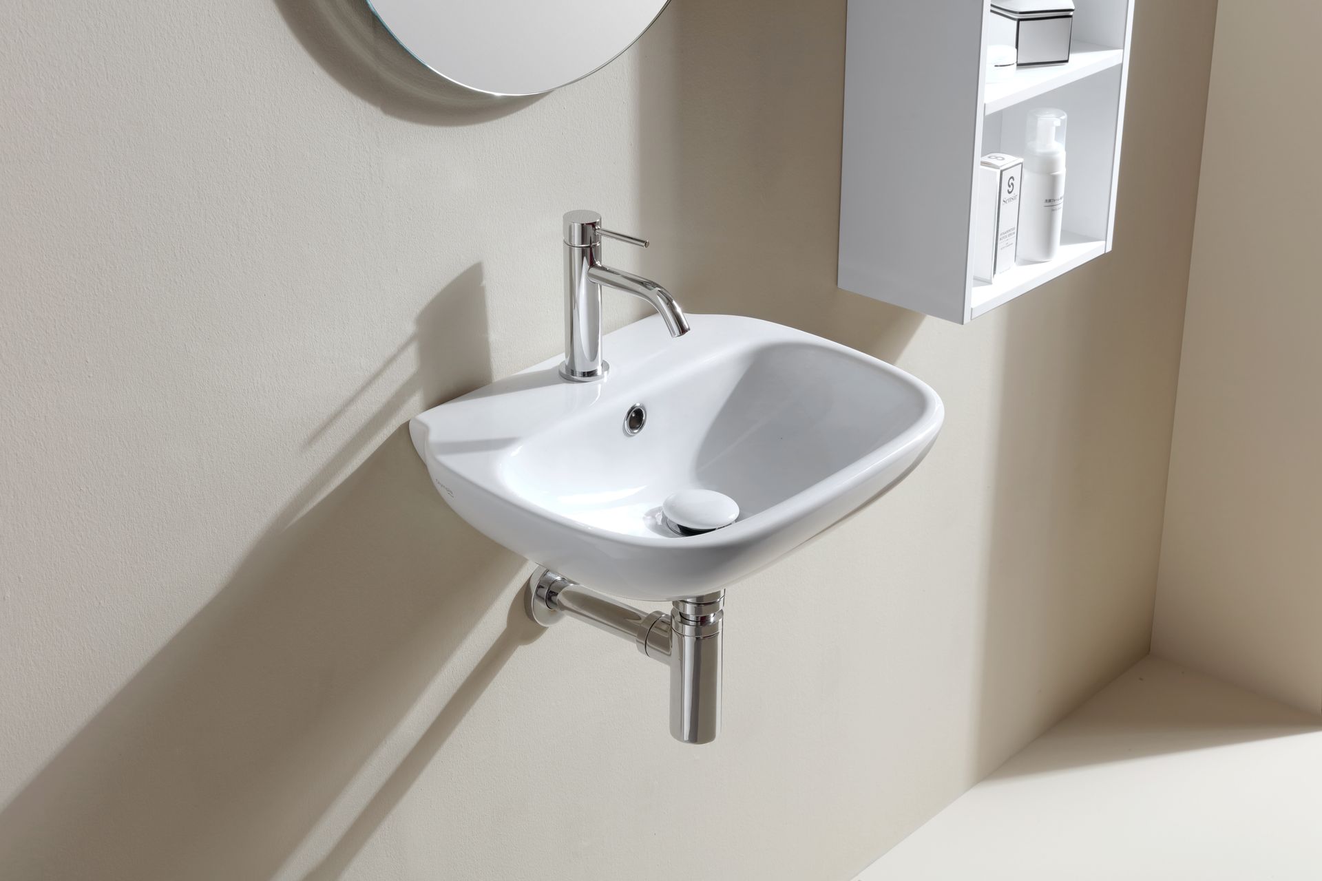 Lavandino in ceramica bianca a parete con rubinetto cromato e tubo di scarico contro una parete beige, con arredo.
