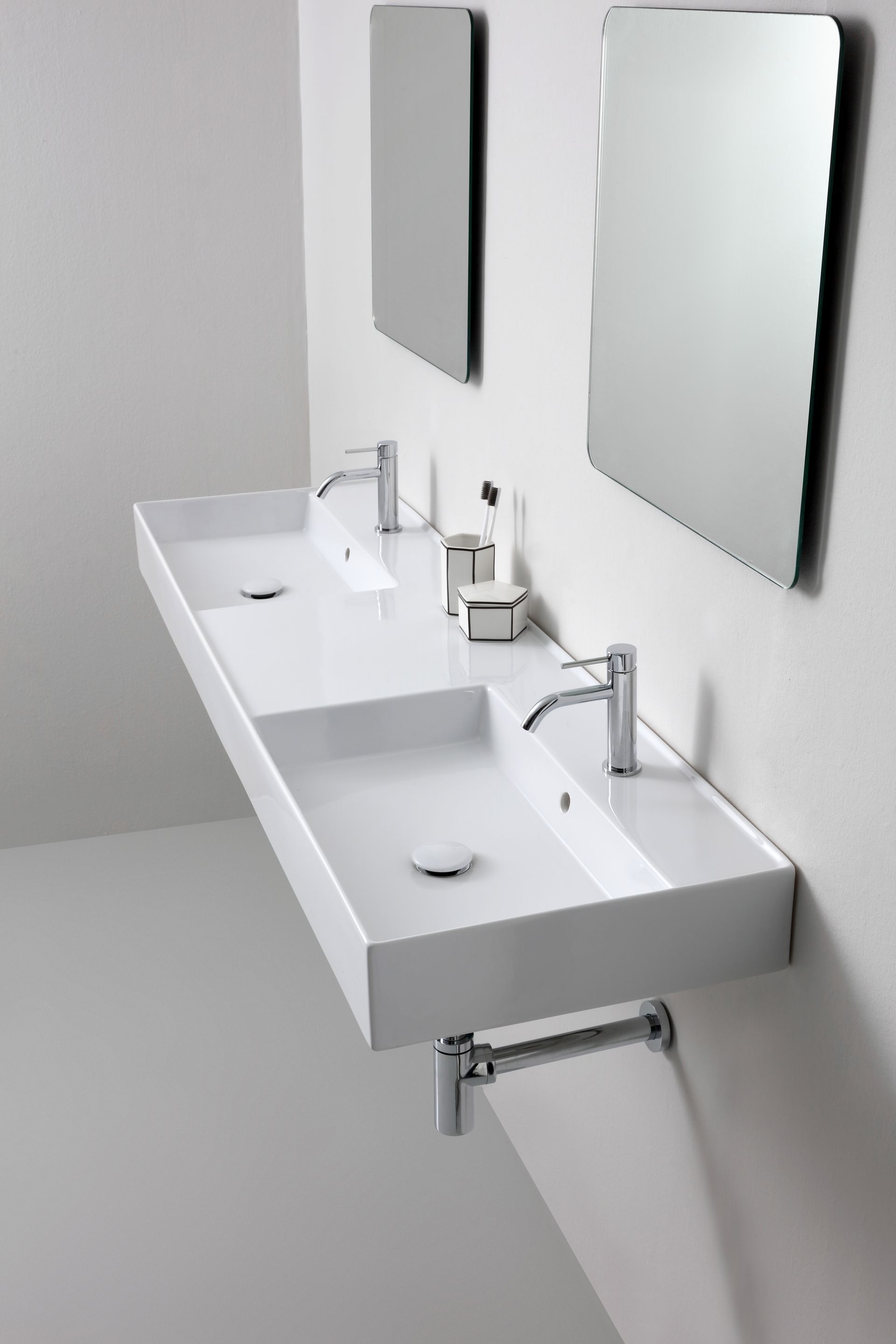Moderno lavabo doppio bianco a parete con due rubinetti e due specchi quadrati, contro una parete bianca.