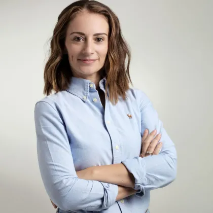 Donna con i capelli castani, camicia blu abbottonata, braccia incrociate, sorriso leggero, su sfondo bianco.
