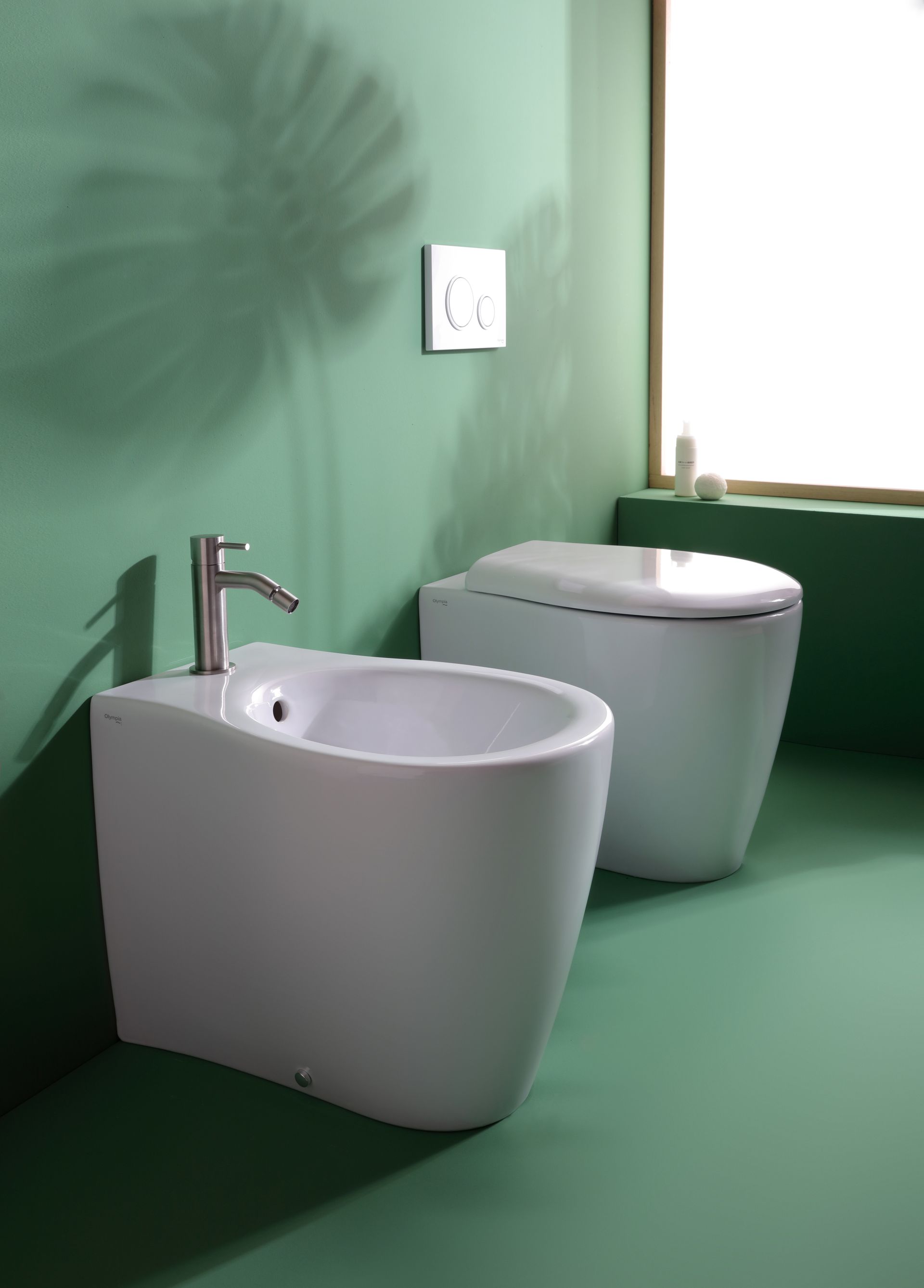 Bidet e WC bianchi in un bagno verde, accanto a una finestra.
