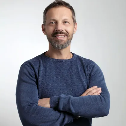 Un uomo con i capelli e la barba brizzolati sorride, con le braccia incrociate e un maglione blu.