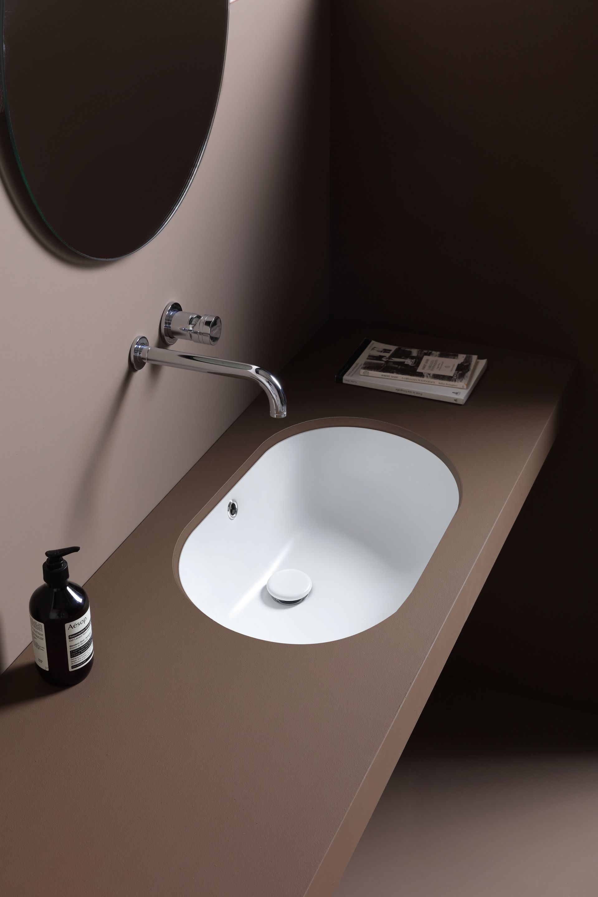 Lavabo da bagno moderno con specchio ovale, rubinetto cromato, dispenser di sapone e asciugamano piegato su un piano scuro.