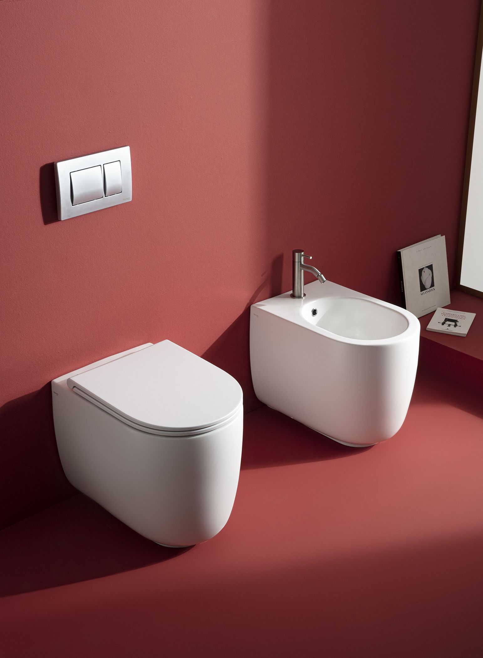 WC e bidet bianchi su un pavimento del bagno rosso, con una placca di scarico argentata sulla parete.