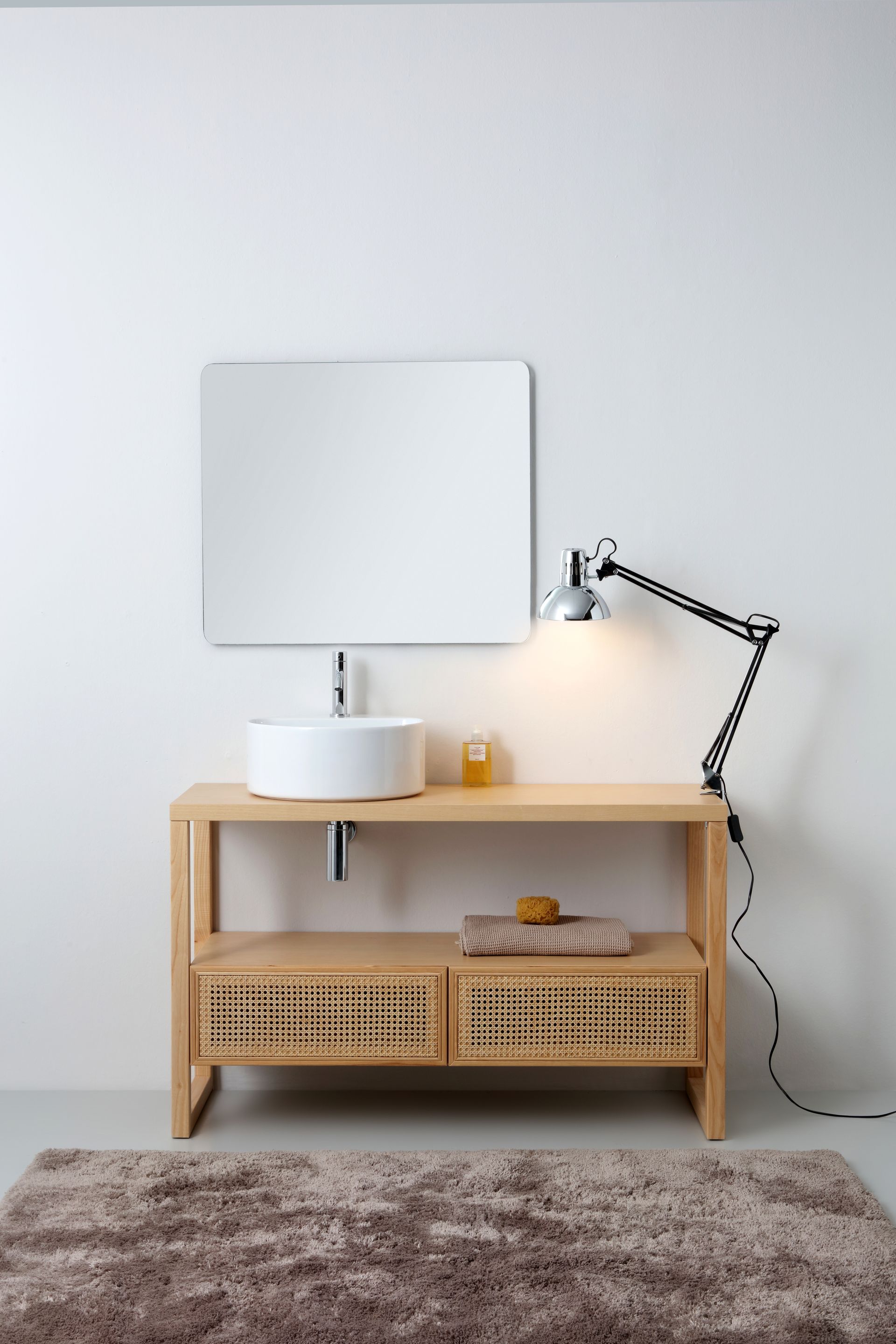 Mobiletto del bagno con lavandino rotondo, specchio e lampada, contro una parete bianca con un tappeto sul pavimento.