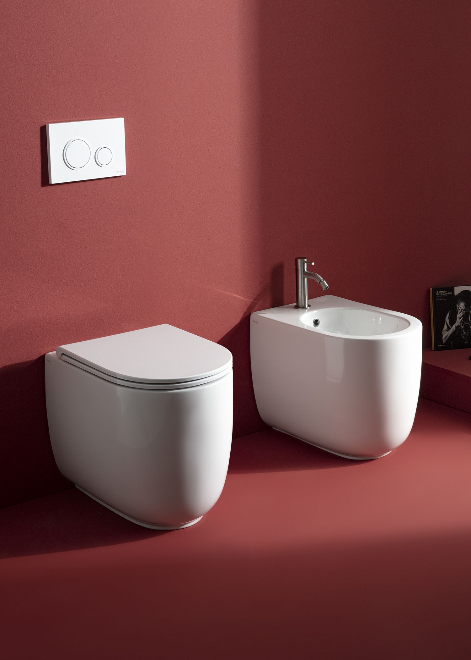 WC e bidet bianchi contro una parete rossa. Rubinetto argentato sul bidet. Comando di scarico bianco.