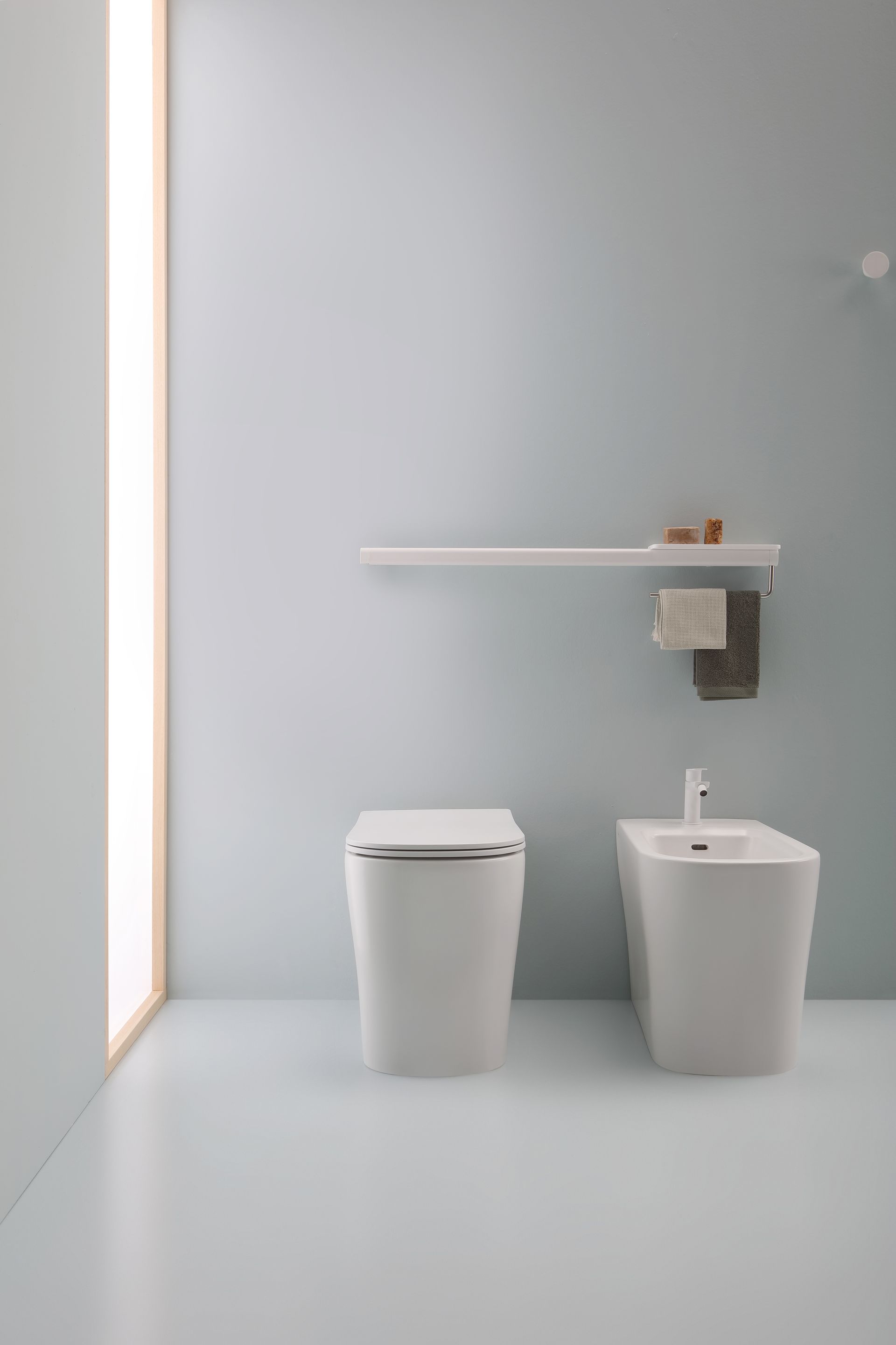 Bagno moderno con WC, bidet e mensola. Pareti e pavimento azzurri. La luce del sole filtra da sinistra.