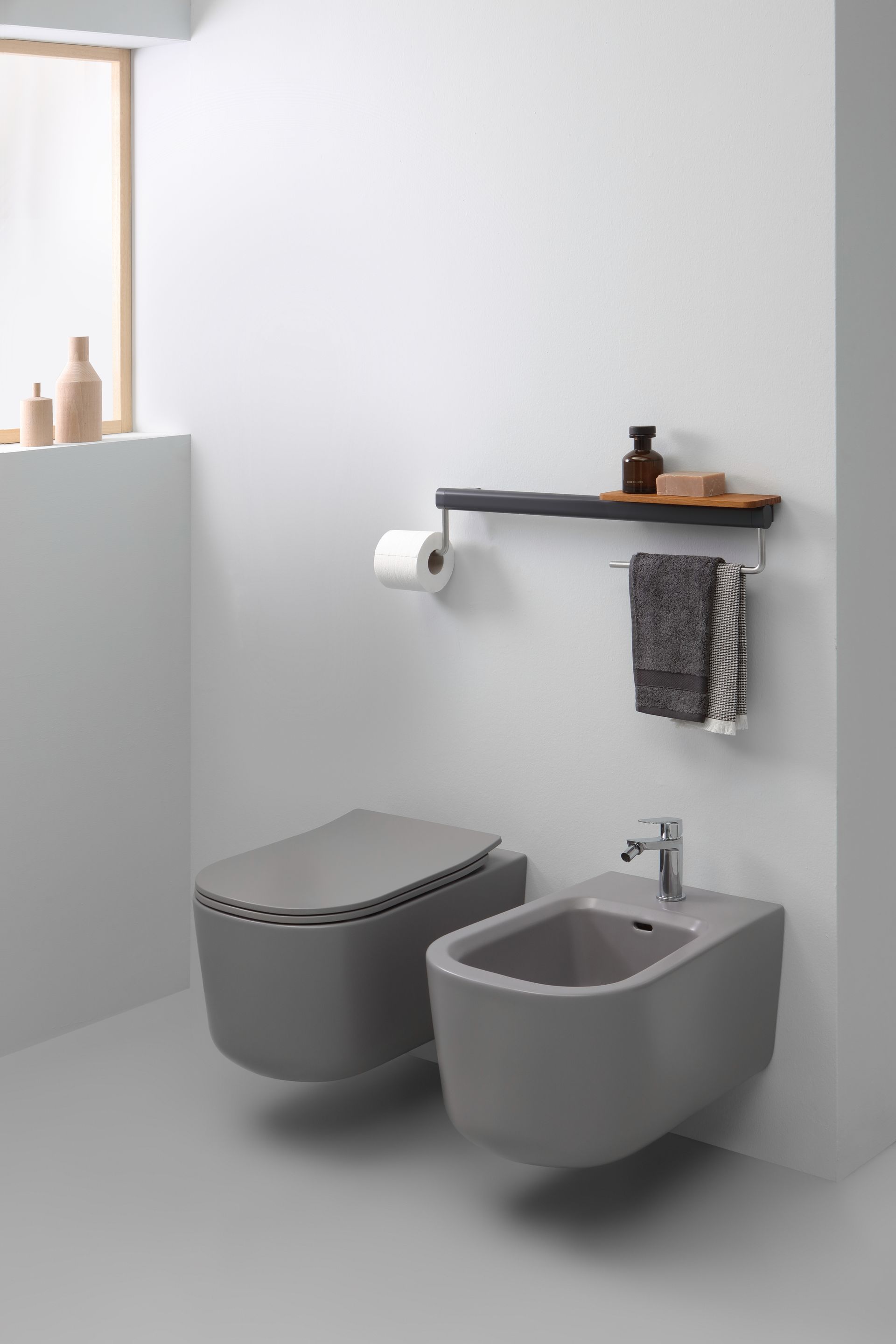 WC e bidet sospesi grigi in un bagno minimalista con mensola, asciugamano e carta igienica.