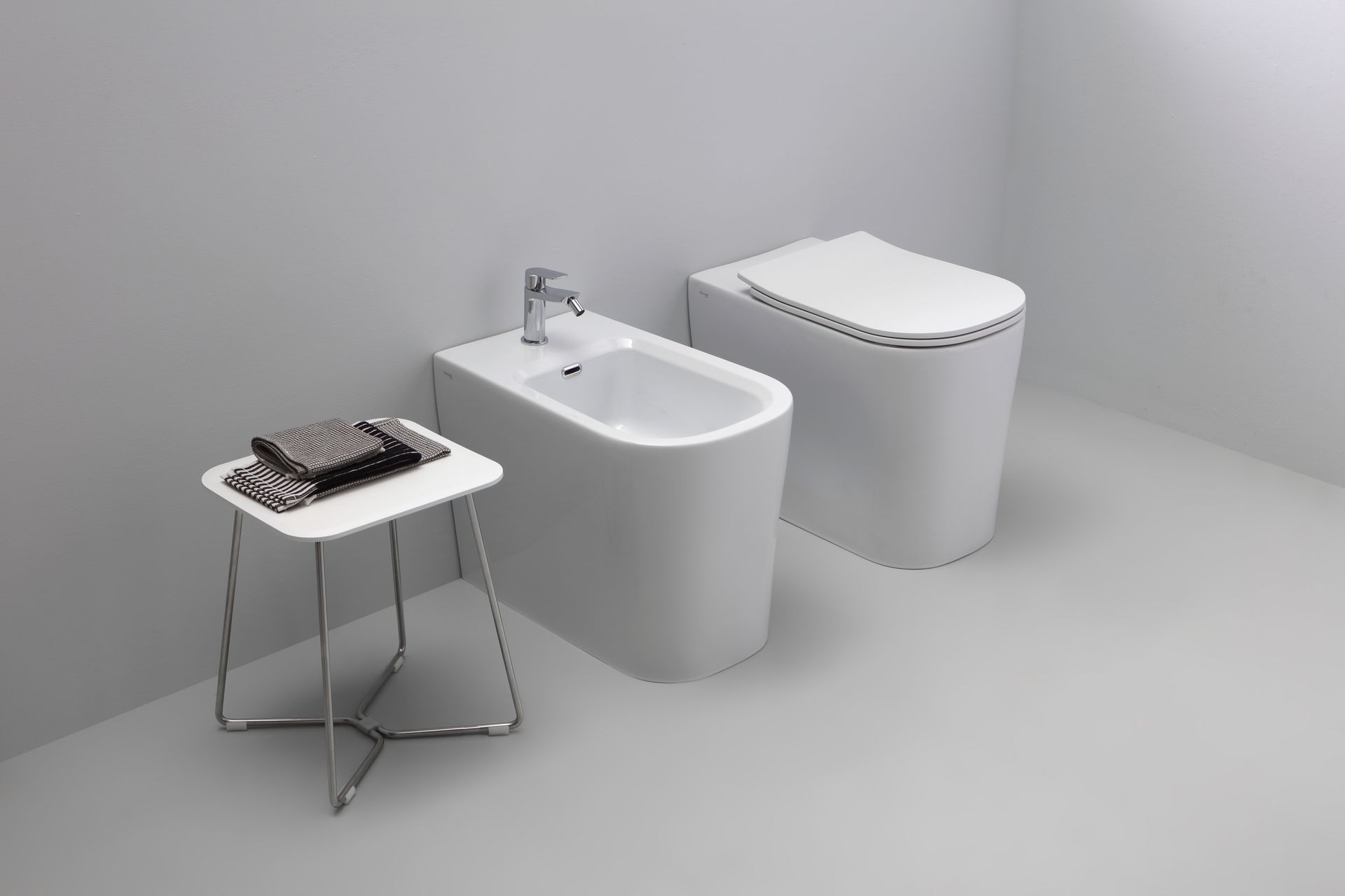 Bagno moderno con WC bianco, bidet e piccolo tavolino con asciugamani su parete e pavimento grigi.