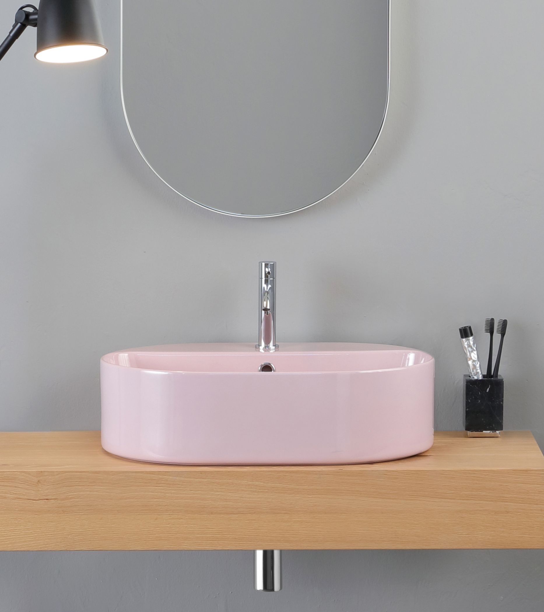 Lavandino rettangolare rosa su mensola in legno, rubinetto argentato, specchio ovale, parete grigia.