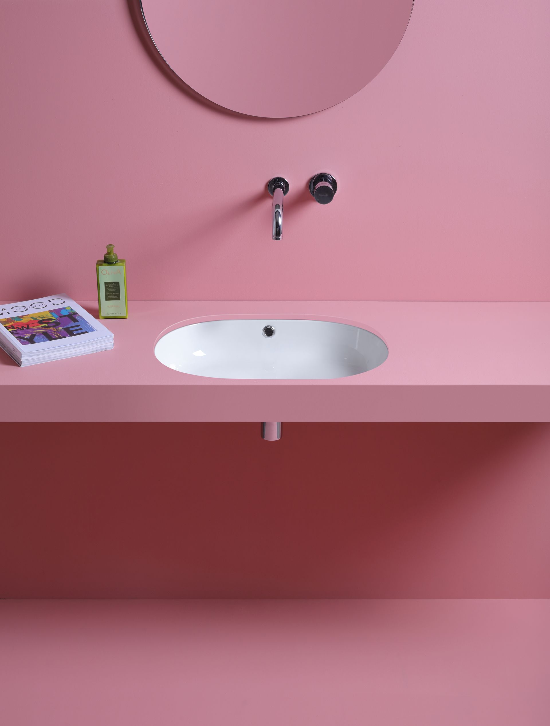 Bagno rosa con lavandino ovale, rubinetto, specchio e ripiano. Sul ripiano ci sono una bottiglia e una rivista.