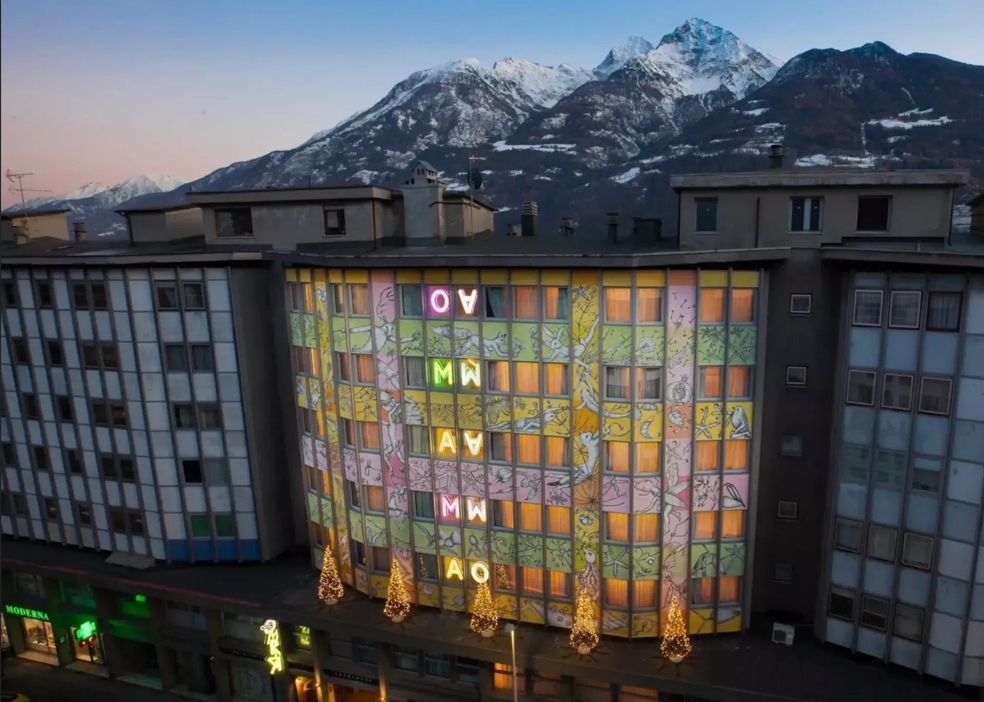 Facciata di un edificio con lettere e finestre illuminate, sullo sfondo delle montagne. Luci colorate e un'atmosfera festosa.