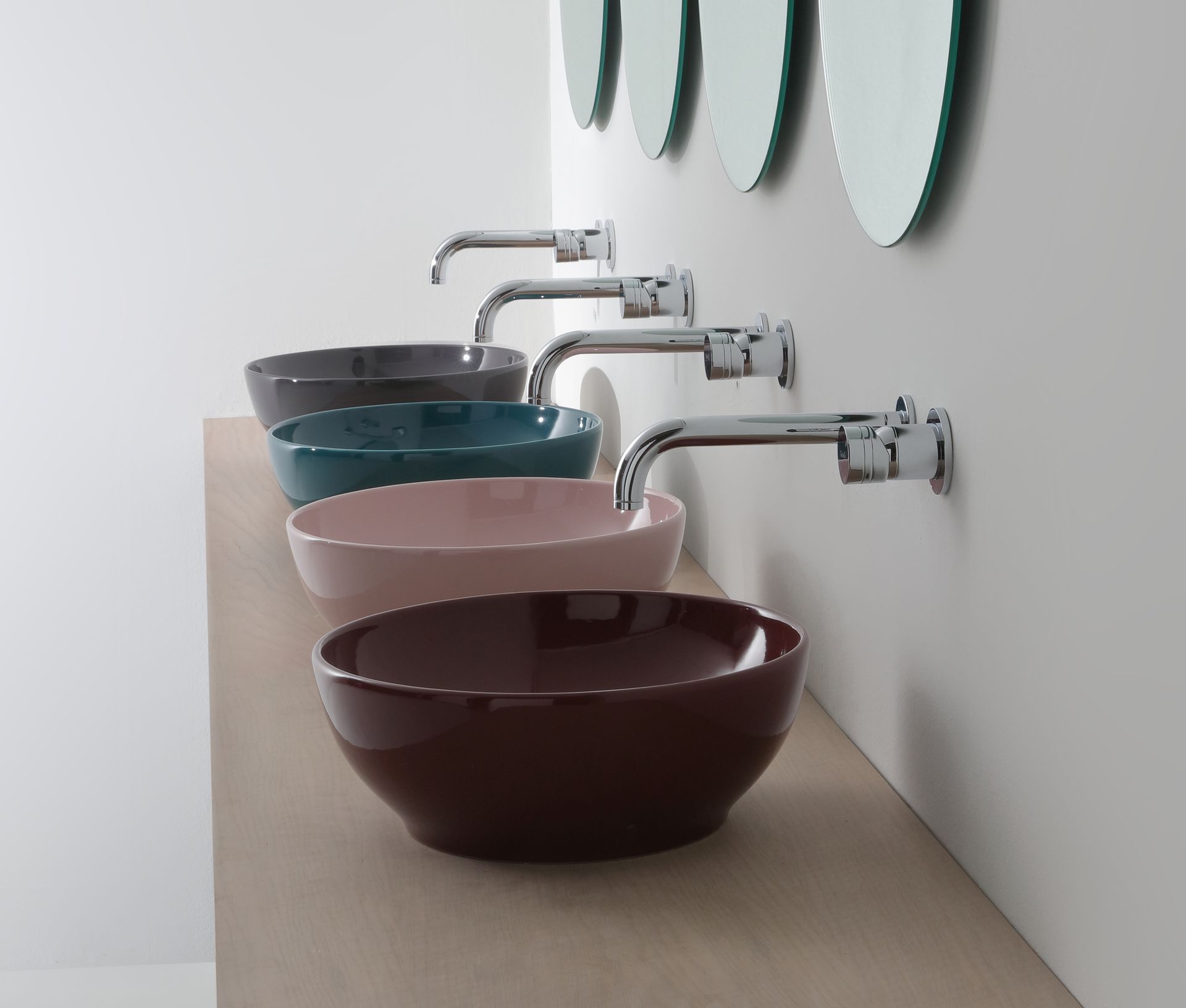 Quattro lavandini colorati con rubinetti a parete su una mensola in legno sotto specchi rotondi.