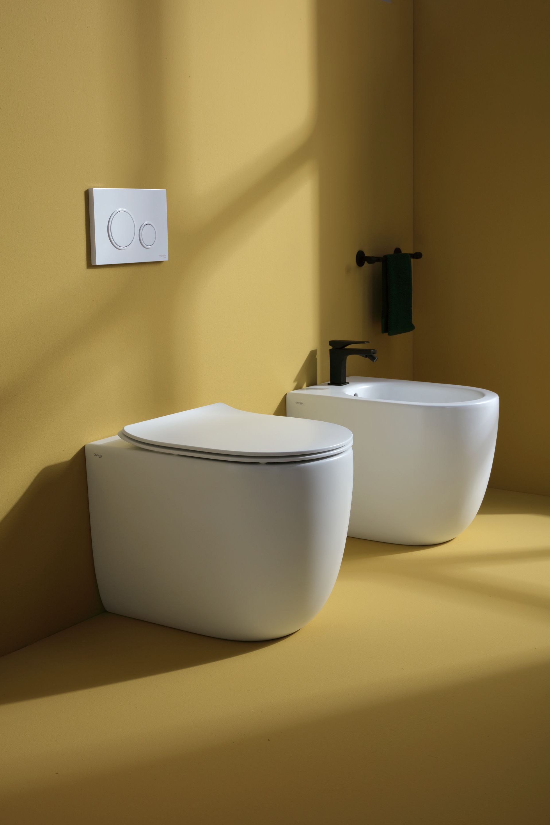 WC e bidet bianchi in un bagno con pareti gialle; sanitari neri, asciugamano verde e pulsante di scarico quadrato.