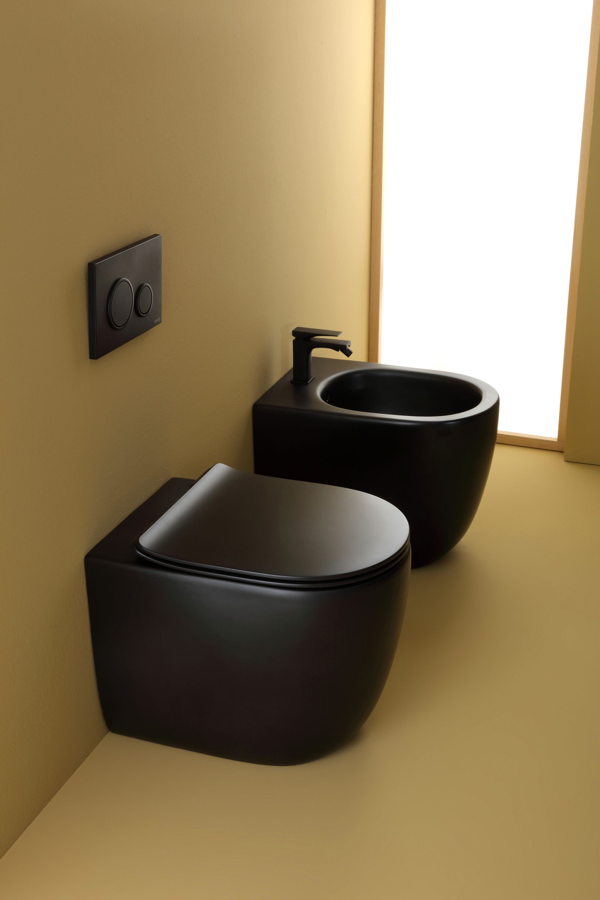 WC e bidet neri in un angolo del bagno, con comando di scarico a parete e finestra.