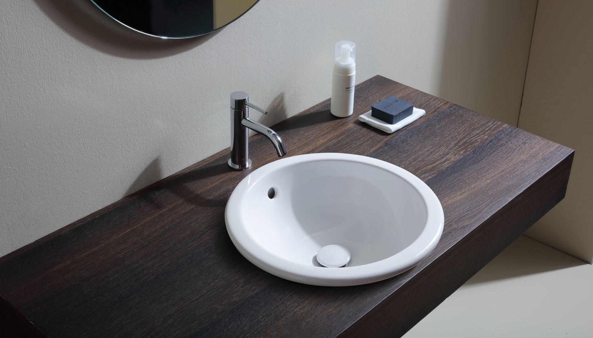 Lavandino rotondo bianco su un piano di lavoro in legno scuro con rubinetto cromato, sapone e bottiglia.