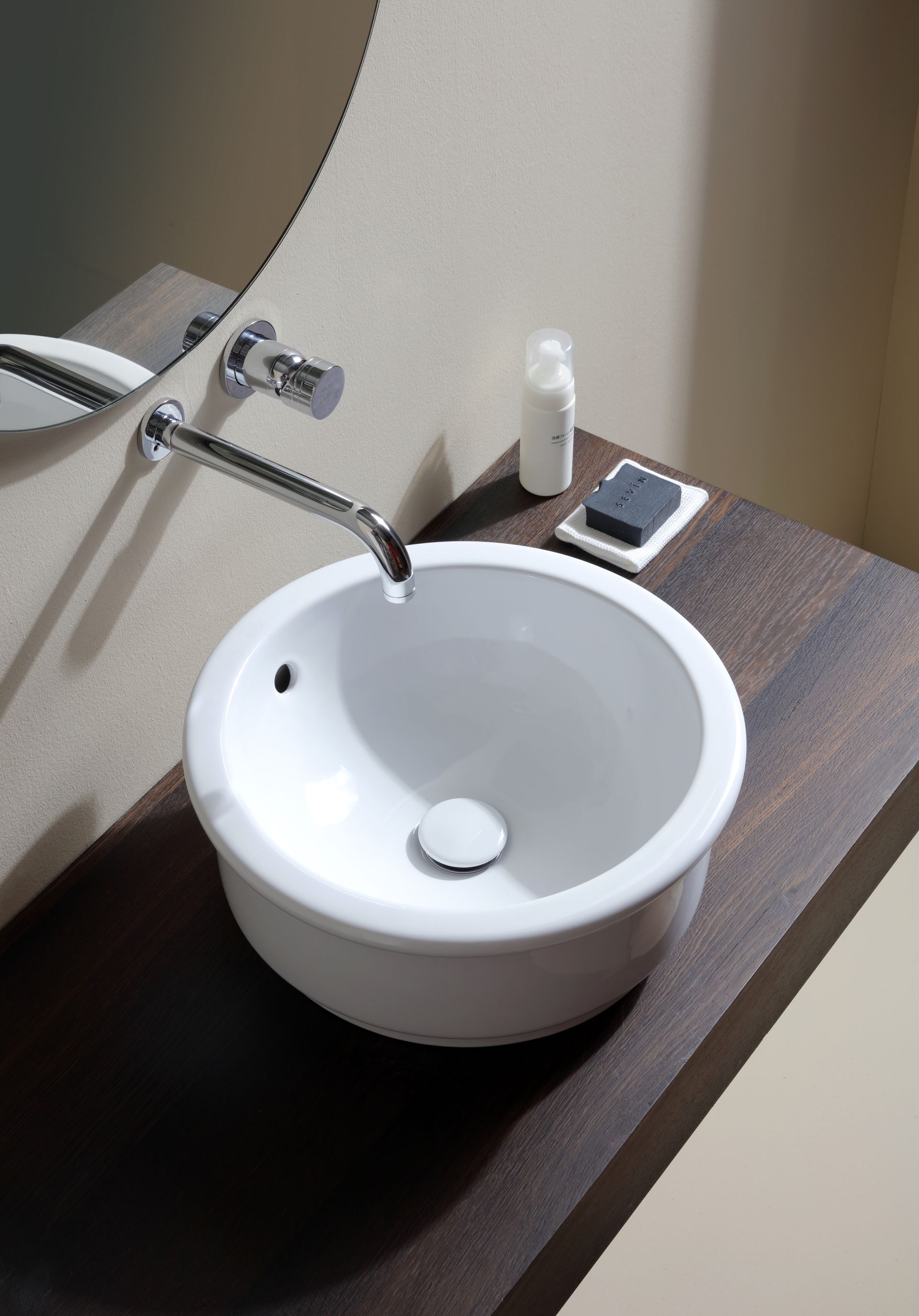 Lavandino bianco rotondo su un piano di lavoro in legno scuro con rubinetto a parete e accessori.
