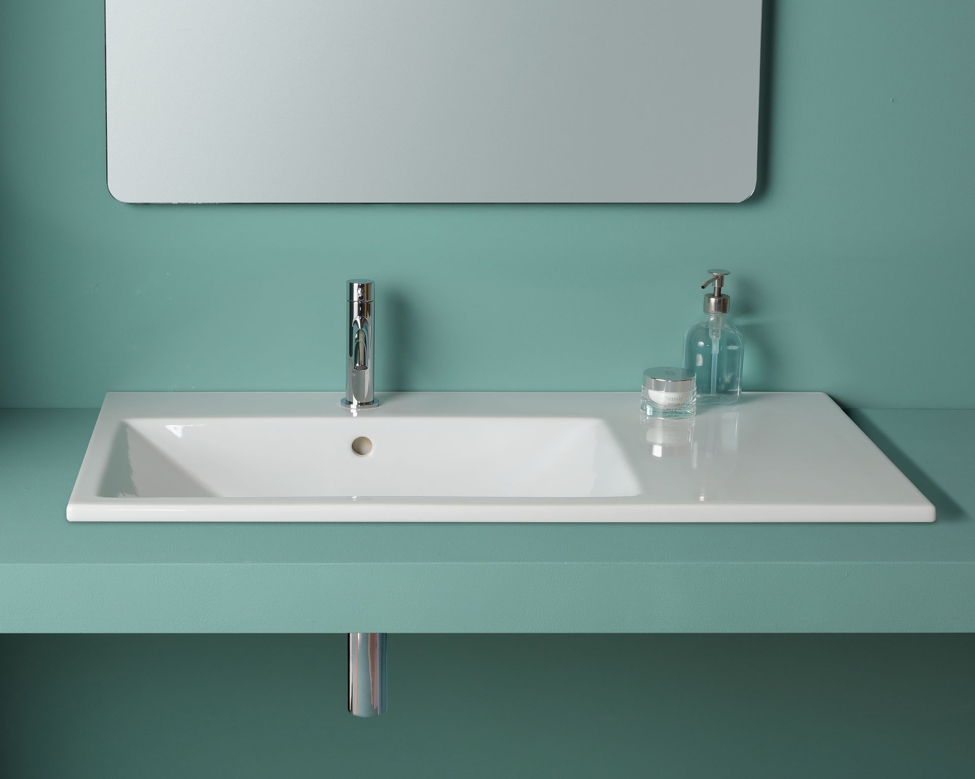 Lavandino rettangolare bianco su un piano di lavoro verde acqua con rubinetto, dispenser di sapone e specchio.