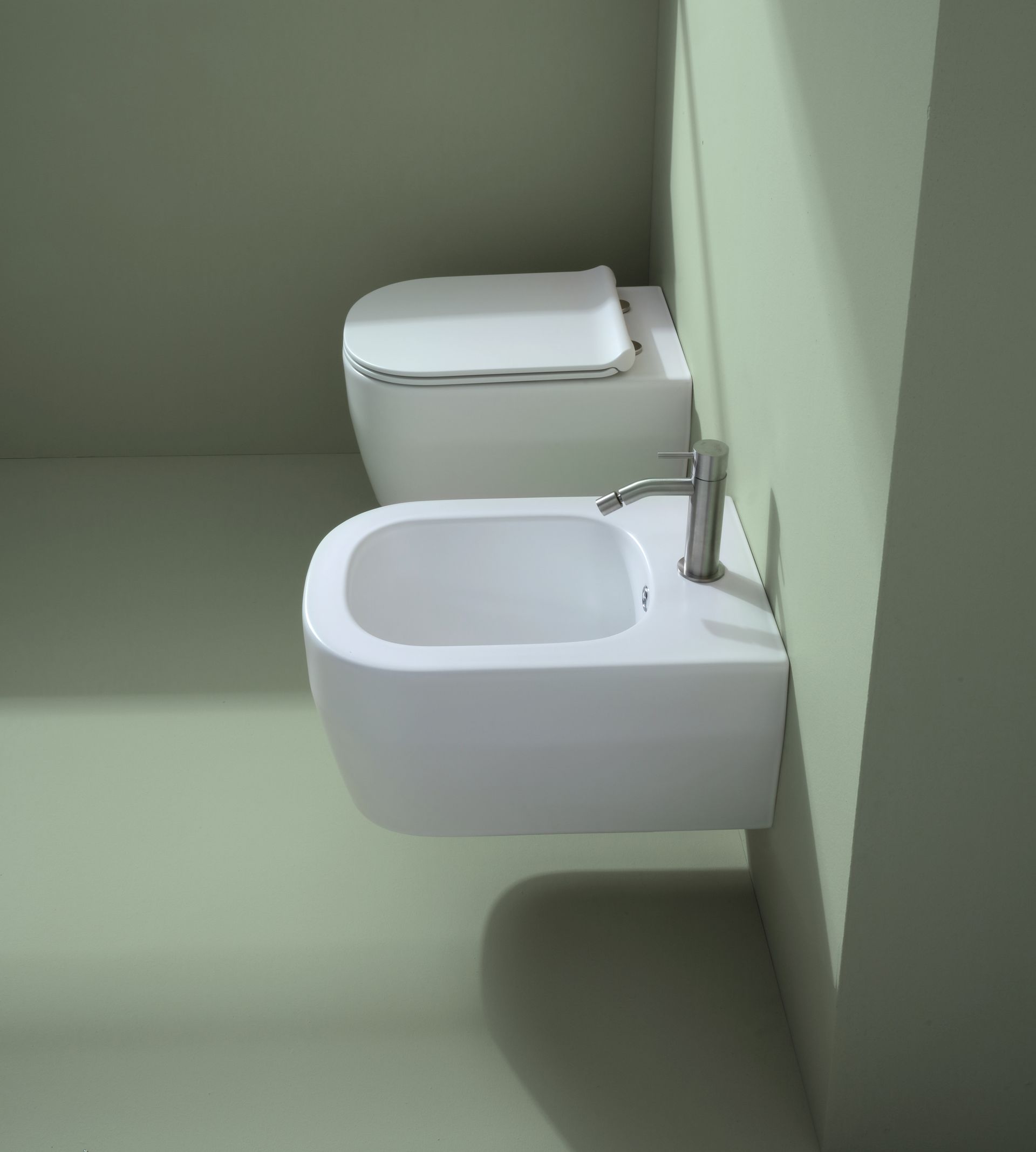WC e bidet bianchi montati su una parete verde, con un rubinetto argentato sul bidet.