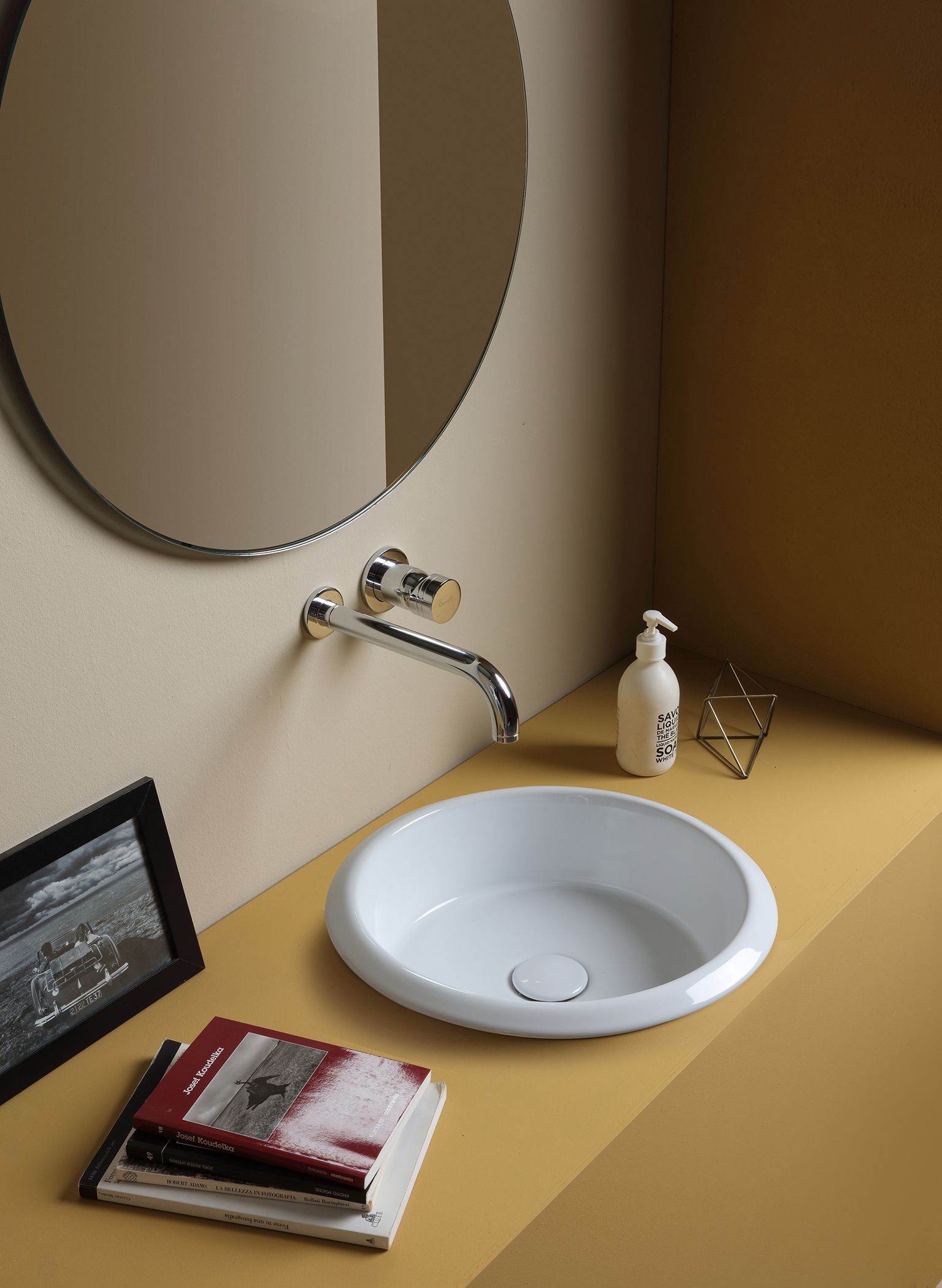 Lavandino bianco con rubinetto in un angolo con pareti gialle, specchio rotondo, dispenser di sapone e libri.