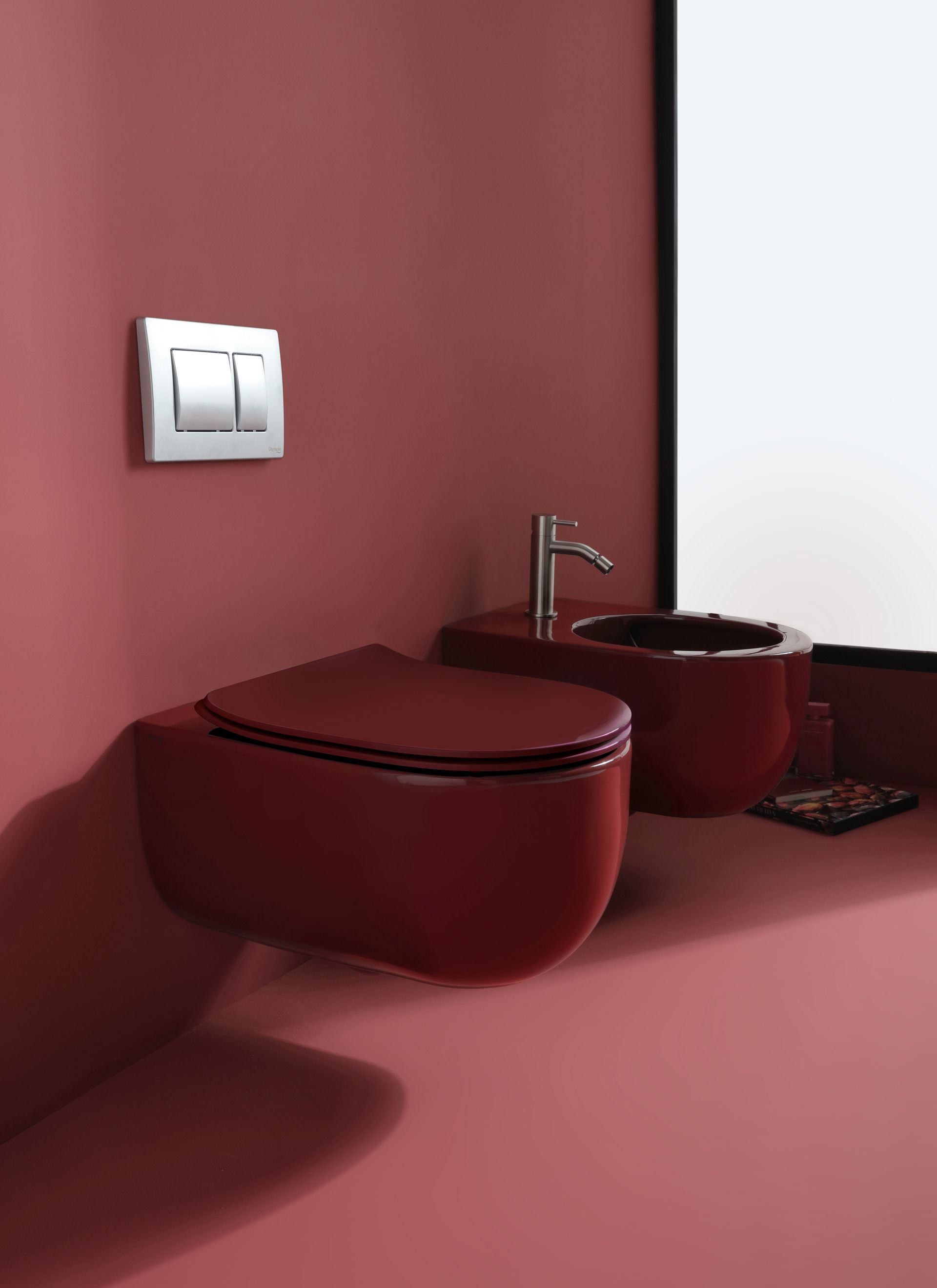 WC e bidet sospesi rossi in un bagno. Placca di scarico cromata. Pavimento e pareti rossi.