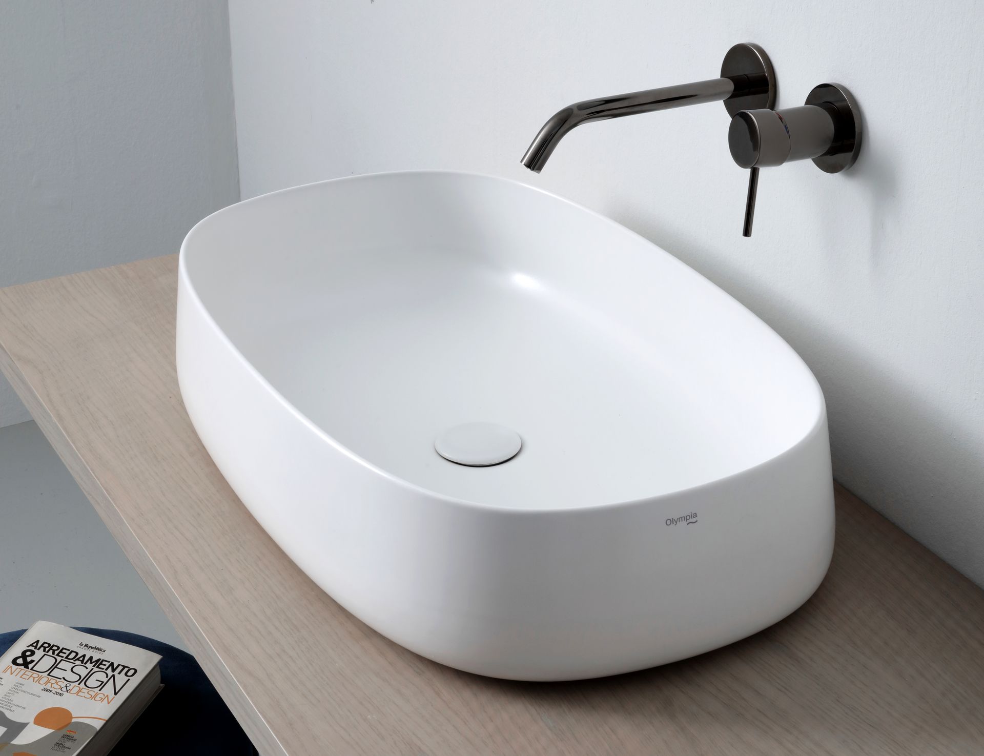 Lavandino ovale bianco su un piano in legno chiaro, rubinetto nero, parete bianca.
