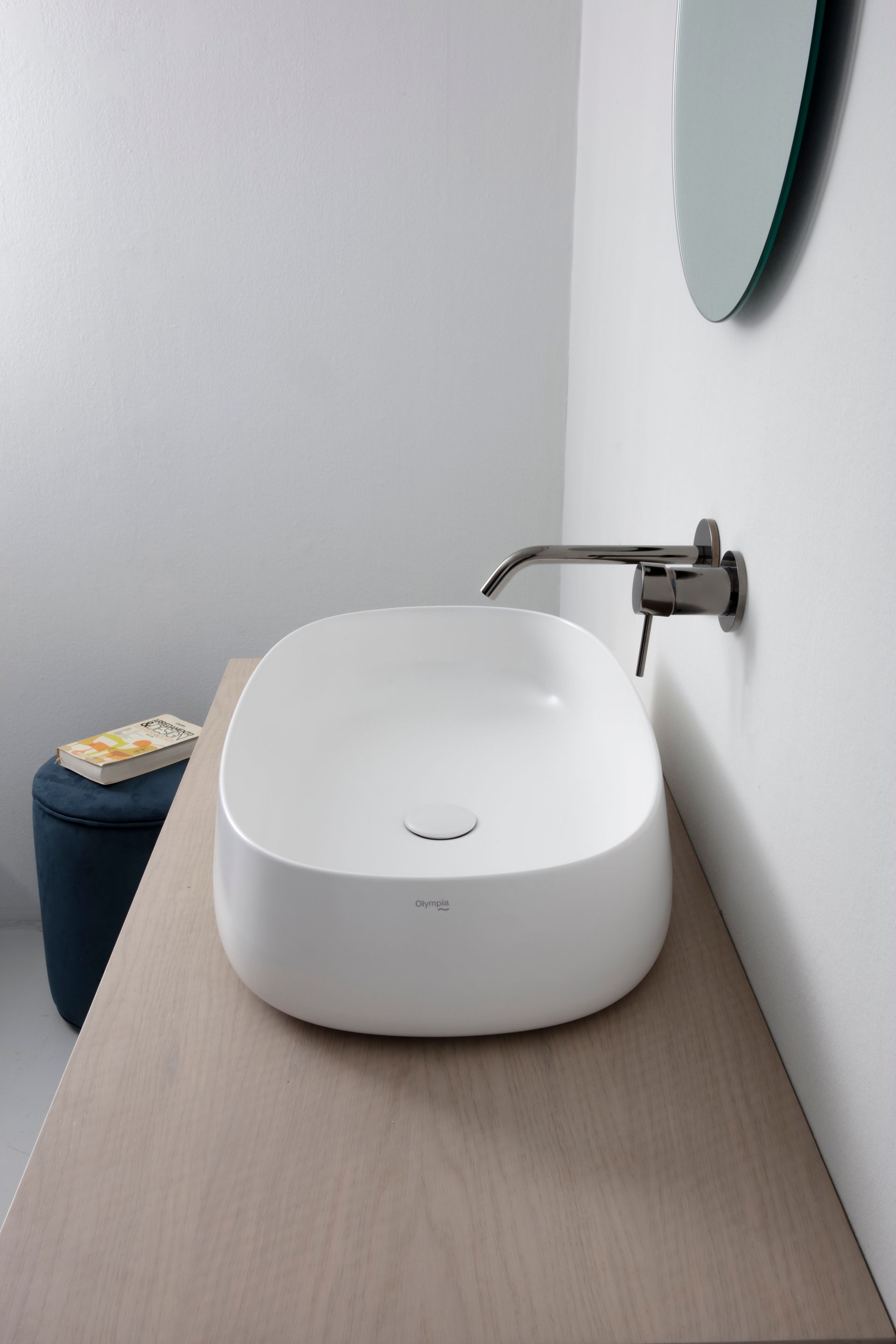 Lavandino ovale bianco su un piano in legno chiaro con rubinetto scuro, pareti bianche e uno sgabello blu.