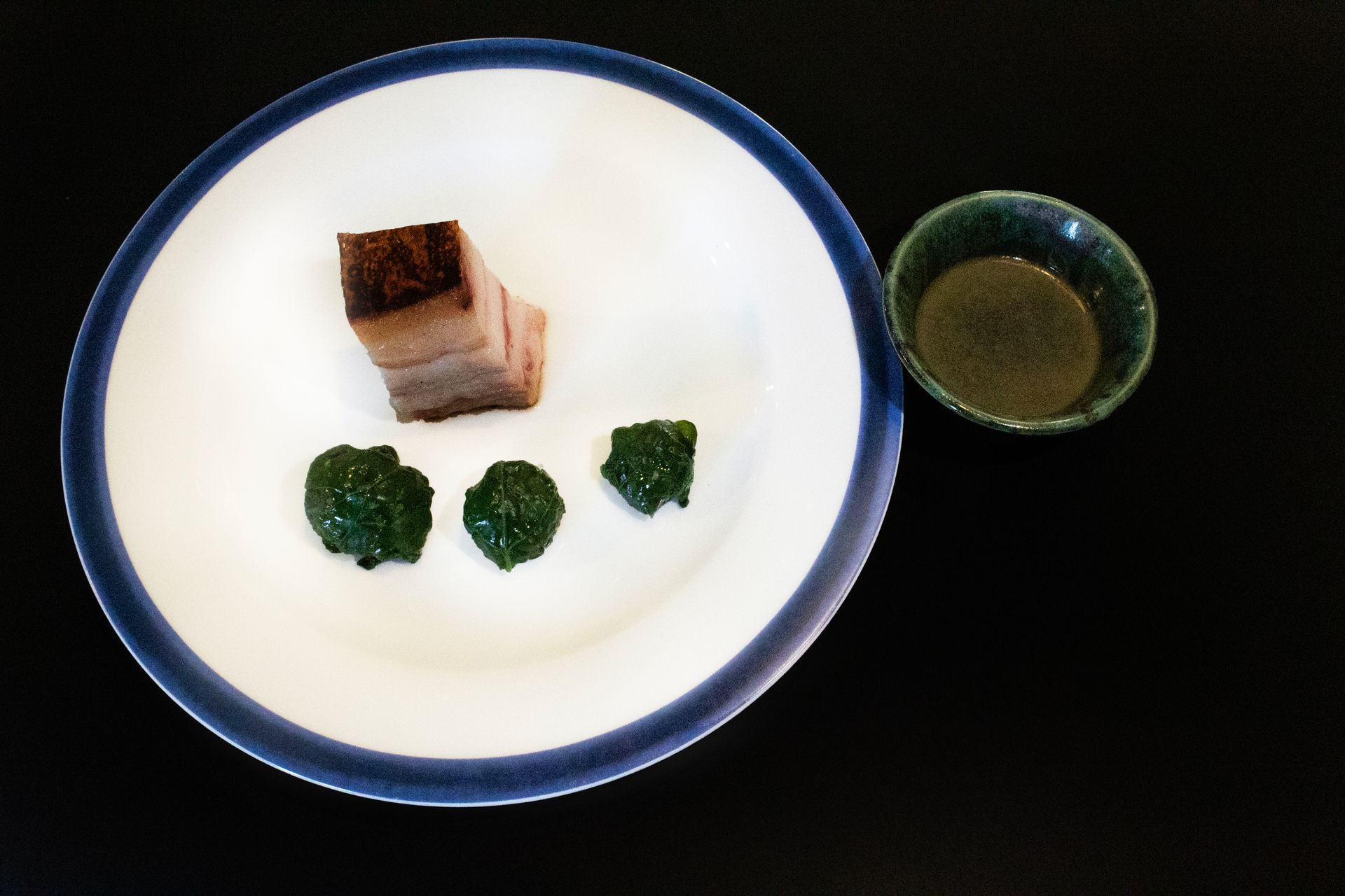 Piatto con tre pezzi di broccoli verdi, un cubetto dorato e una ciotolina di salsa su sfondo nero.