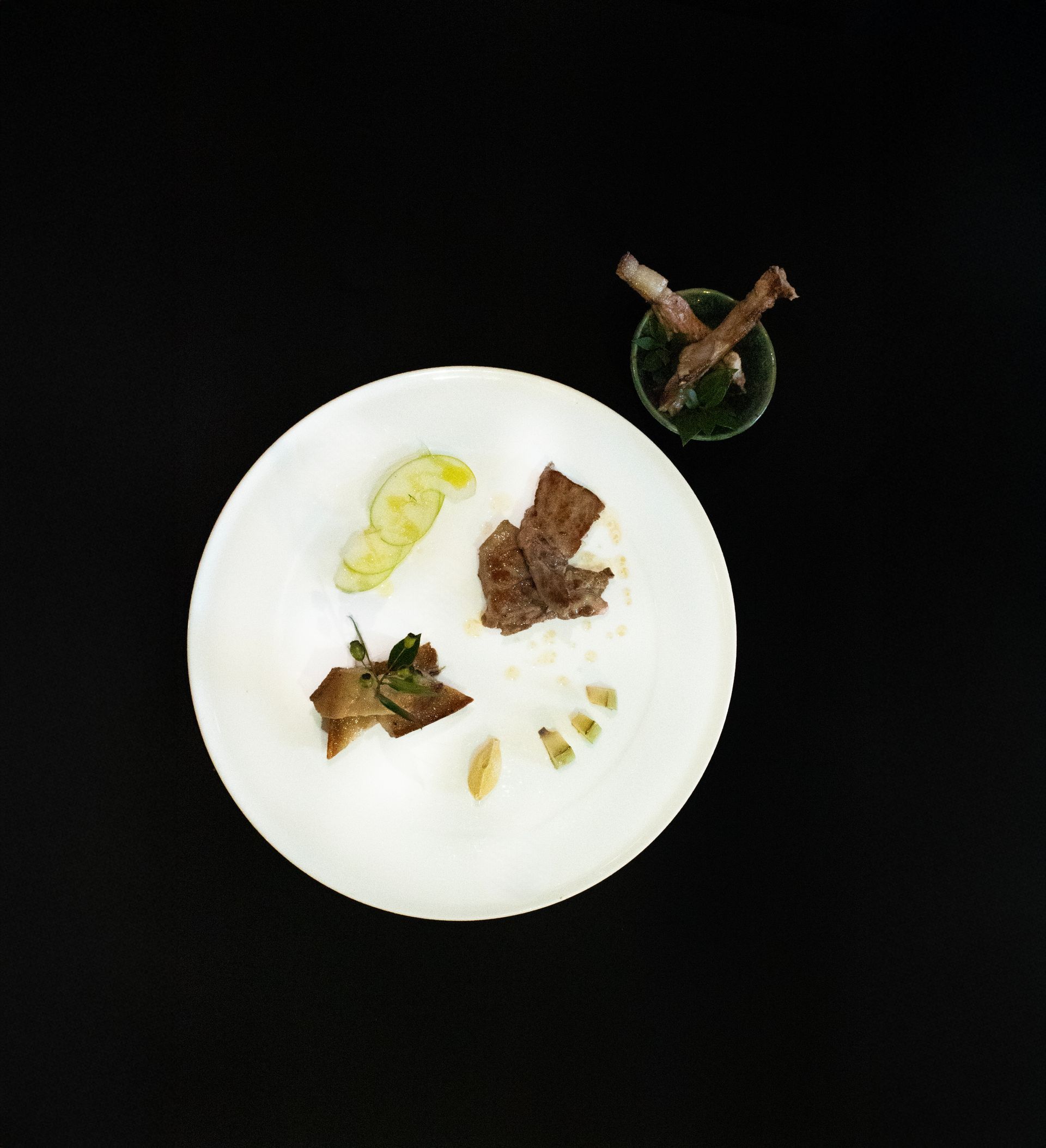 Vista dall'alto di un piatto bianco con diverse piccole porzioni di cibo su un tavolo scuro, più un piccolo bicchiere sulla destra.