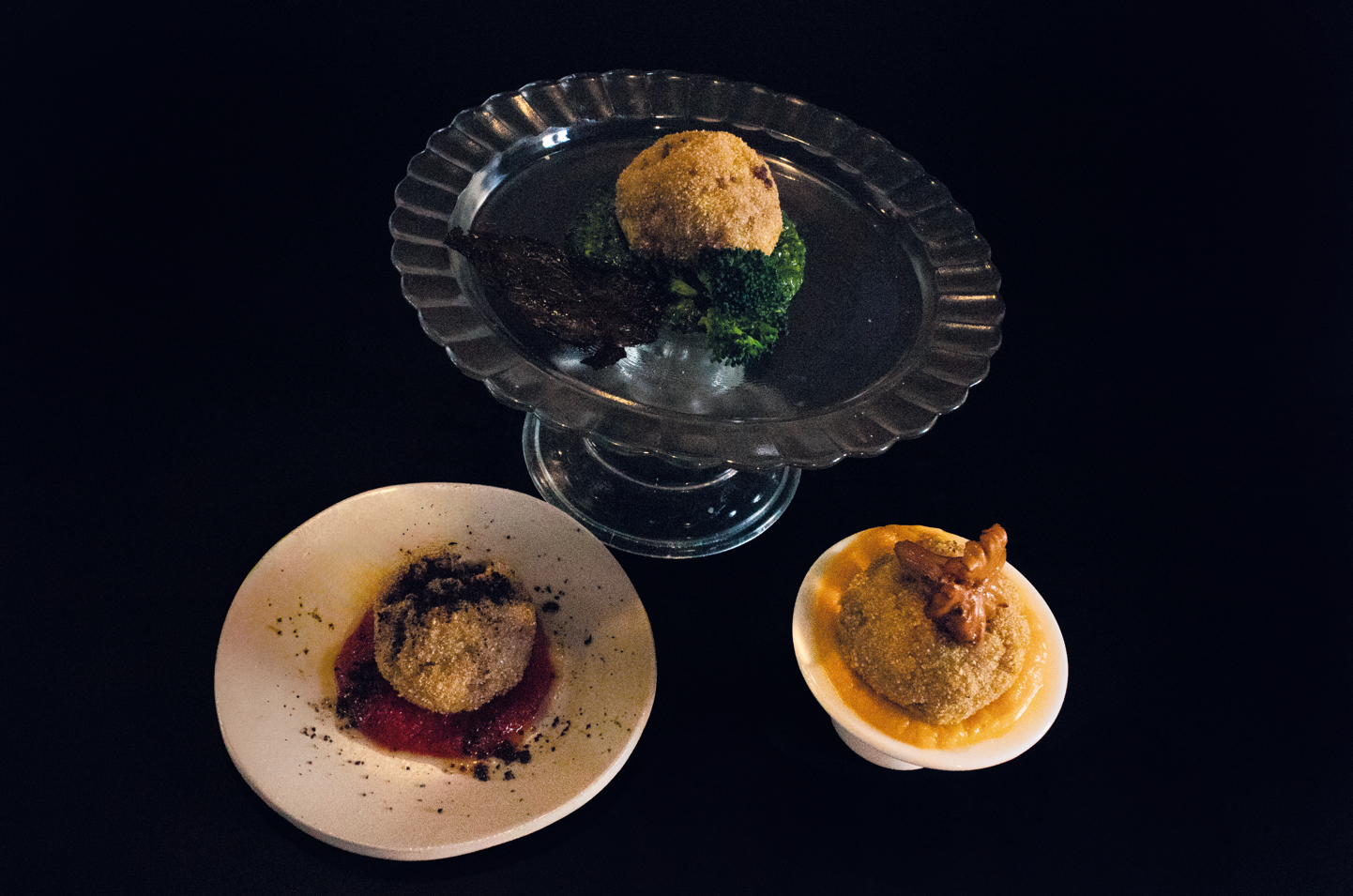 Tre dessert impiattati su sfondo nero, tra cui un piatto di vetro con piedistallo e due piattini bianchi.