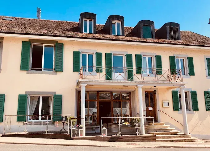 Un immeuble couleur crème avec des volets verts, un balcon et une entrée centrale vitrée sous un ciel bleu.