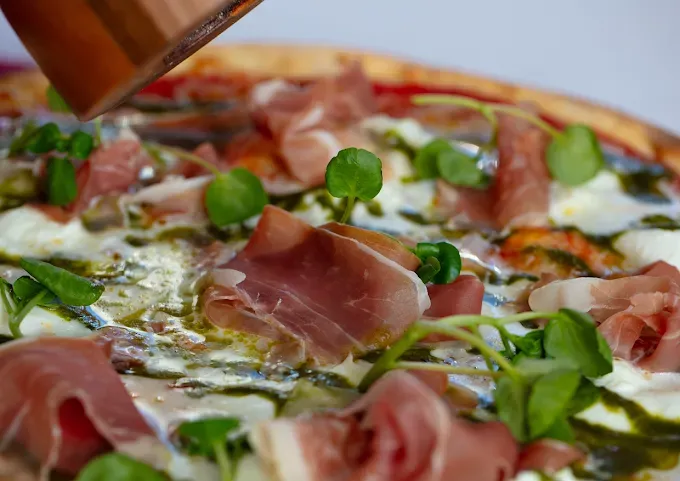 Gros plan sur une pizza garnie de prosciutto, de salade et de fromage fondu, en cours de découpe.
