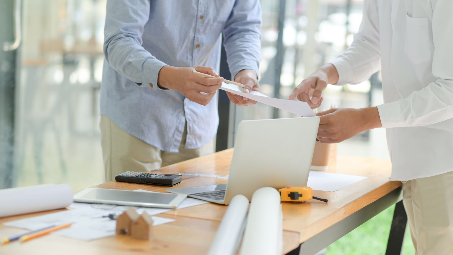 Two men in an office reviewing papers over a laptop on a desk with blueprints and a small model house.