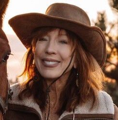 Woman wearing a cowboy hat smiles, standing next to a horse outdoors.