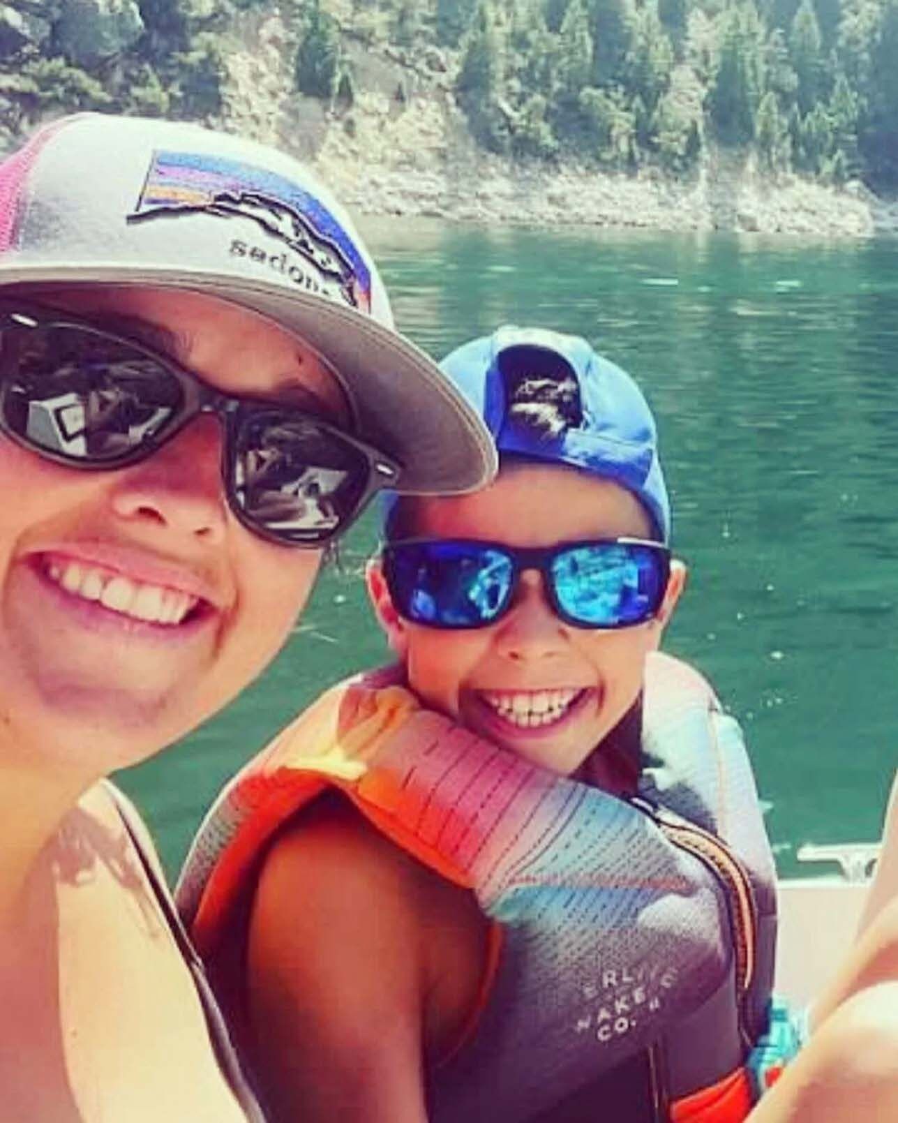 Woman and child smiling on a boat, wearing sunglasses and life vests, with a lake and trees in the background.
