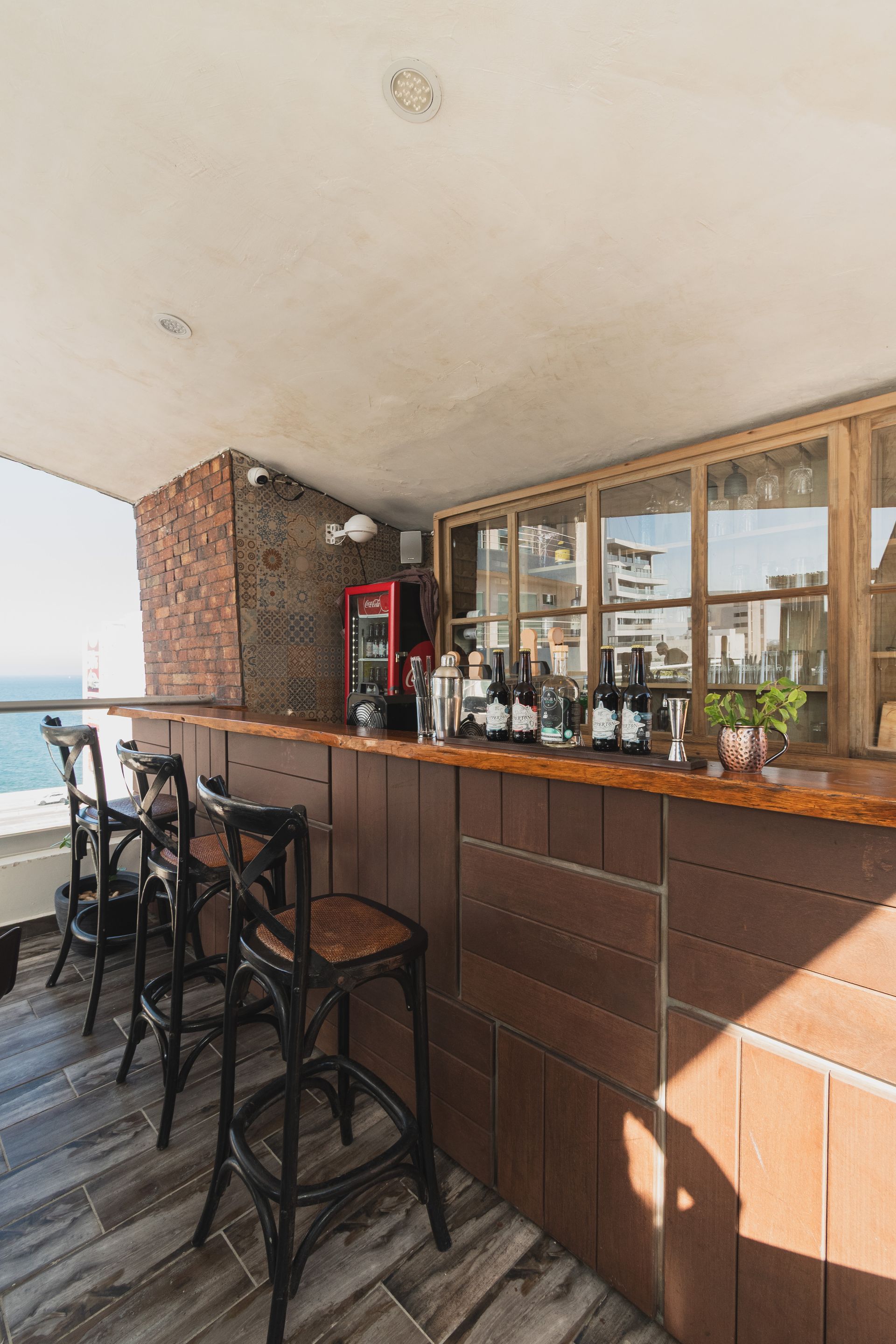 A rustic outdoor bar with wooden paneling, tall dark chairs, and a glass-paneled display case overlooking the ocean.