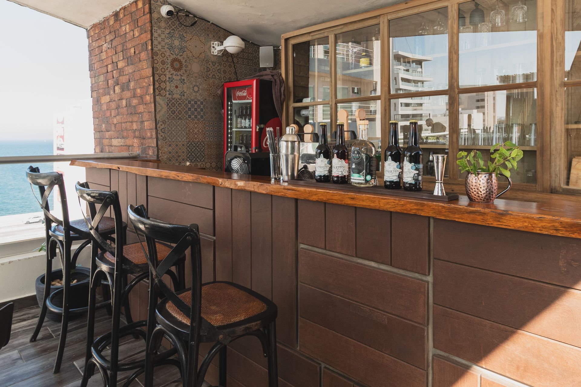 Bar rústico al aire libre con barra de madera y sillas negras, situado junto a una pared de ladrillos con vista al océano