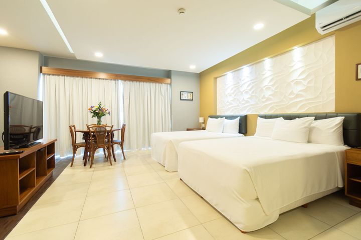 Habitación de hotel con dos camas blancas, una mesa de madera con sillas, un televisor y grandes cortinas blancas.