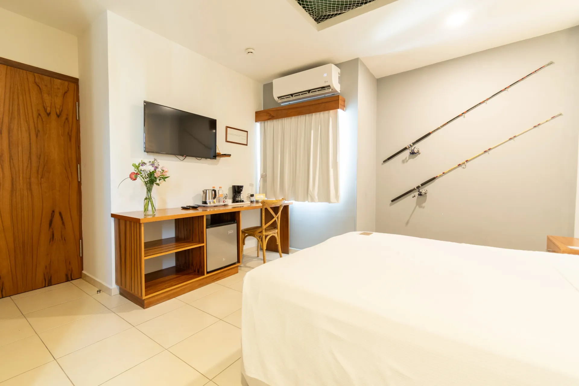 A hotel room with a bed, a television, a desk with a small refrigerator, and two fishing rods mounted on the wall.