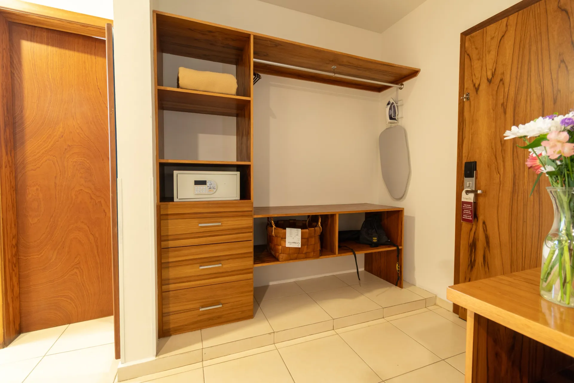 A wood-paneled closet area with shelves, a safe, drawers, a hanging rack, ironing board, and a nearby floral arrangement.