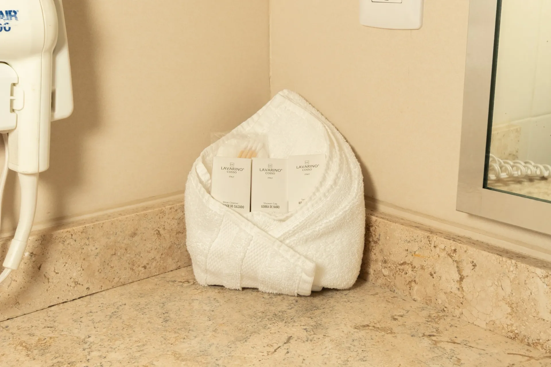 A white folded towel holding small toiletry bottles on a beige marble hotel bathroom counter.