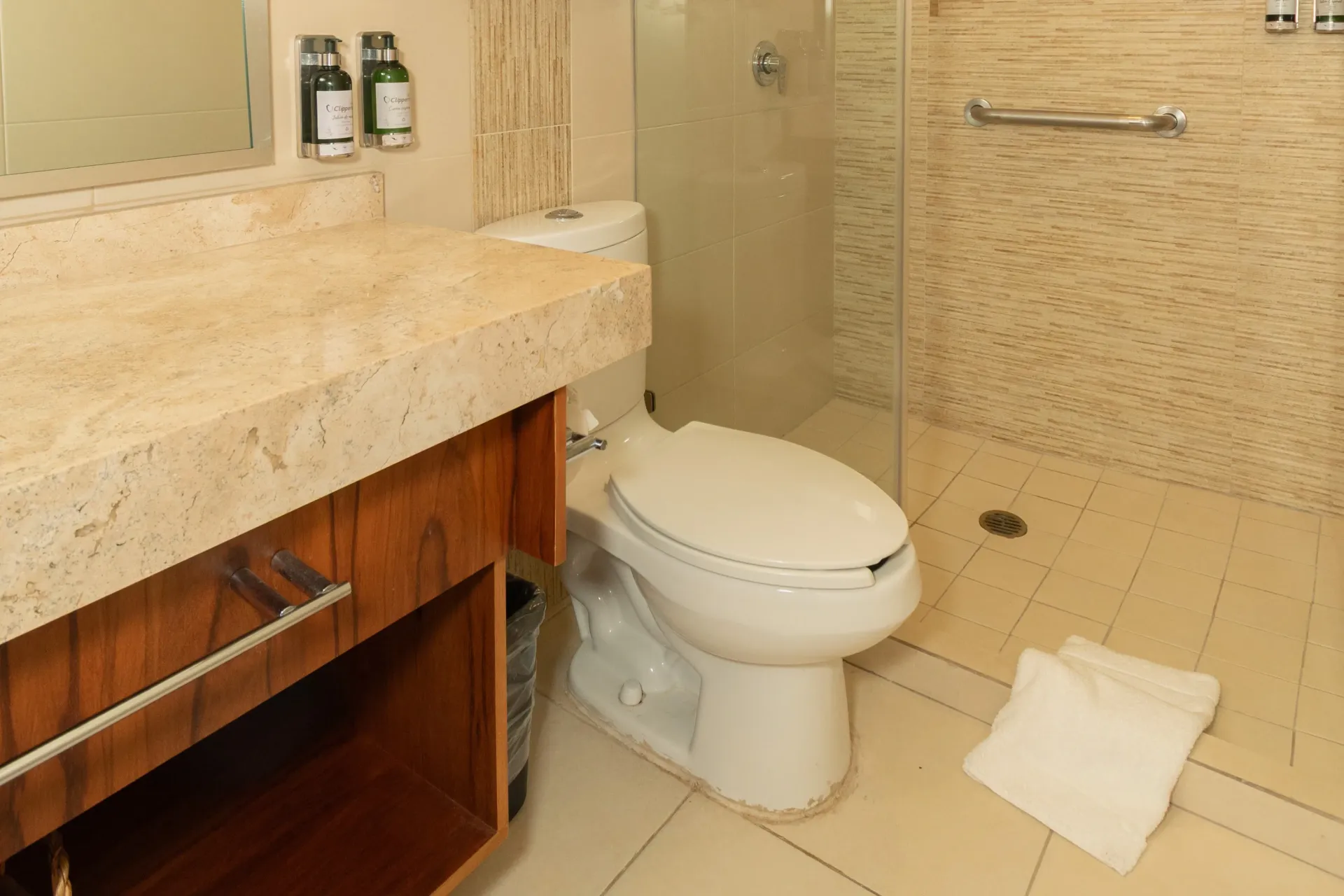A bathroom with a beige stone vanity, a white toilet, and a glass-enclosed shower with a handrail.